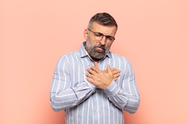 Homem adulto bonito parecendo triste, magoado e com o coração partido, segurando as duas mãos perto do coração, chorando e se sentindo deprimido