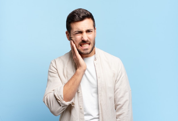 Homem adulto bonito segurando a bochecha e sofrendo de uma dor de dente dolorosa, sentindo-se doente, miserável e infeliz, procurando um dentista