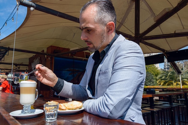 Homem adulto caucasiano de etnia francesa, vestindo roupas elegantes, sentado tomando café da manhã, tomando café com sanduíche, sentado no bar do lado de fora do restaurante