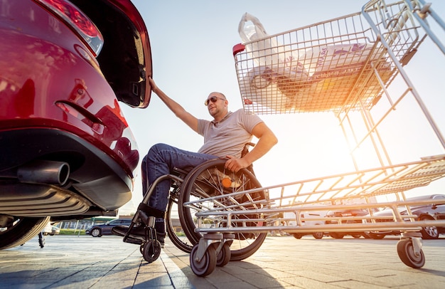 Homem adulto com deficiência em cadeira de rodas coloca as compras no porta-malas de um carro no estacionamento de um supermercado