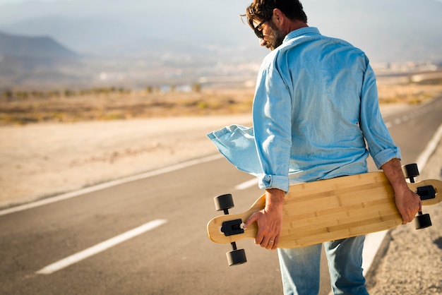 Homem adulto com mesa de prancha longa caminhando em uma longa rua de asfalto - conceito de pessoas modernas alternativas e transporte gratuito para viagens - pessoas ativas ao ar livre