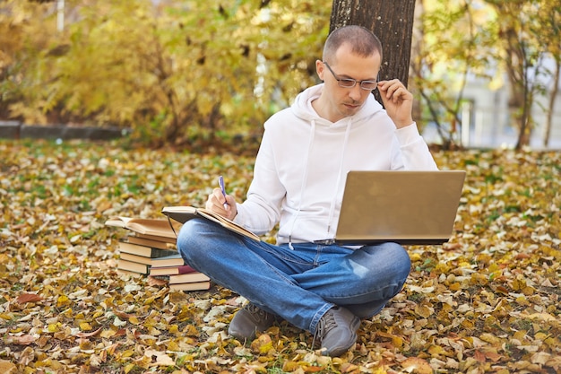Homem adulto com um moletom branco está estudando no parque em um laptop, escrevendo em um caderno, lendo livros e livros didáticos. aprendizagem ao ar livre, distanciamento social