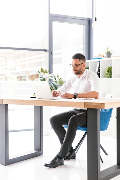 Homem adulto de 30 anos, bem sucedido, de corpo inteiro, com roupa formal e óculos, sentado à mesa e usando um laptop, enquanto trabalha na sala de escritório