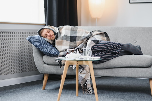 Homem adulto doente que tem um vírus de resfriado ou gripe sazonal está lutando contra doenças em casa Infeliz homem de meia idade está sentado no sofá embrulhado em cachecol quente e manta segurando lenço de papel e espirrando