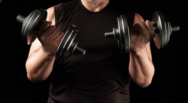 Homem adulto em roupas pretas tem halteres de aço nas mãos, seus músculos estão tensos