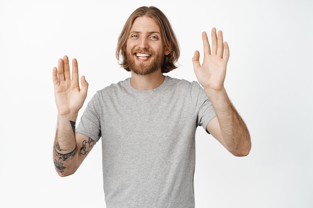 Homem adulto feliz e bonito, loiro acenando com as mãos, gesto de adeus, cumprimentando você, sorrindo amigavelmente, parado no branco