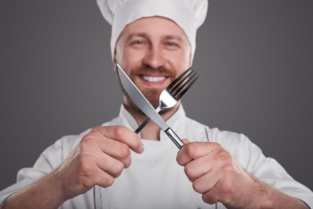 Foto homem adulto feliz em uniforme de chef cruzando garfo e faca e olhando para a câmera com um sorriso durante o trabalho contra um fundo cinza