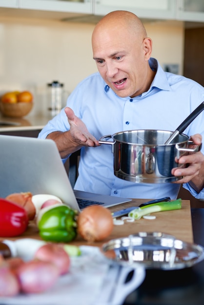 Homem adulto forte na cozinha preparando comida e olhando para o laptop