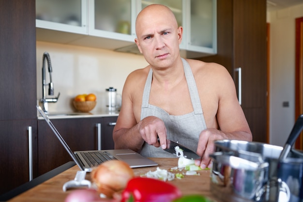 Homem adulto forte na cozinha preparando comida e olhando para o laptop