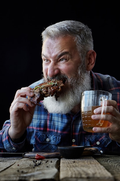 Homem adulto grisalho brutal com barba come o bife de mostarda e bebe cerveja, convida para uma refeição, o conceito de um feriado, festival, Oktoberfest