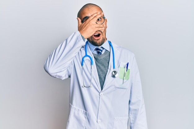 Homem adulto hispânico vestindo uniforme médico e estetoscópio espreitando em choque cobrindo o rosto e os olhos com a mão olhando por entre os dedos com expressão envergonhada