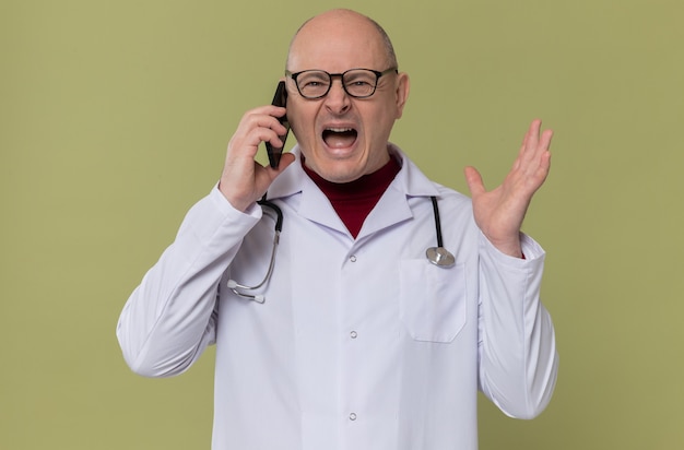 Homem adulto irritado com óculos em uniforme de médico com estetoscópio gritando com alguém no telefone