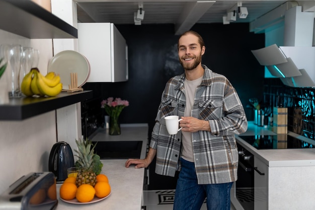 Homem adulto jovem sorridente com barba segurando xícara com café de pé junto ao balcão da cozinha com interior leve moderno desfrutando de bebida quente fresca em casa