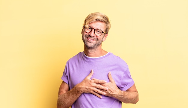 Homem adulto loiro bonito se sentindo romântico, feliz e apaixonado, sorrindo alegremente e segurando as mãos perto do coração