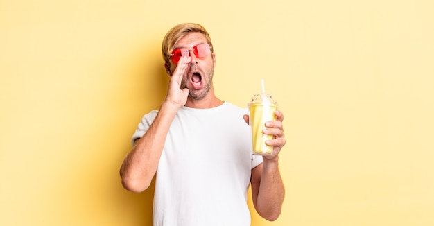 Homem adulto loiro se sentindo feliz, dando um grande grito com as mãos perto da boca com um milkshake