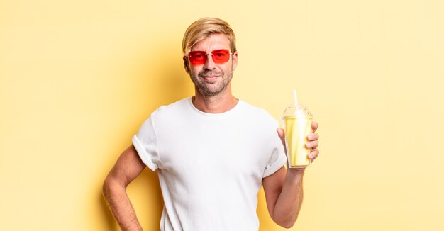 homem adulto loiro sorrindo feliz com uma mão no quadril e confiante com um milkshake