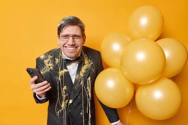 Homem adulto muito feliz vestido com roupas formais manchadas com spray serpantina usa celular para conversar on-line detém um monte de balões inflados isolados sobre fundo amarelo Conceito de férias