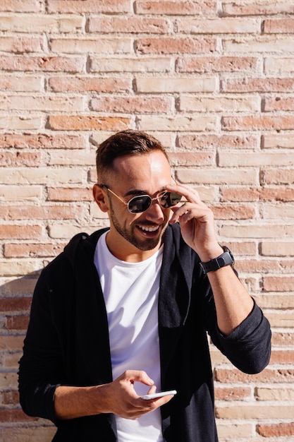 Foto homem adulto sorridente usando óculos de sol usando telefone inteligente enquanto está de pé contra uma parede de tijolos