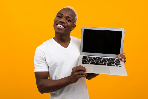 Homem Africano bonito com sorriso bonito mantém computador portátil sem fio com simulação sobre fundo amarelo