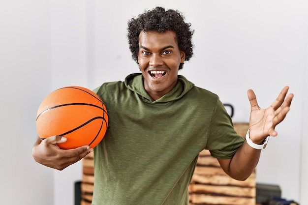 Homem africano com cabelo encaracolado segurando uma bola de basquete no ginásio comemorando a vitória com sorriso feliz e expressão de vencedor com as mãos levantadas