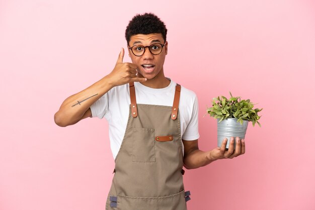 Homem africano de jardineiro segurando uma planta isolada no fundo rosa, fazendo gesto de telefone. ligue-me de volta sinal