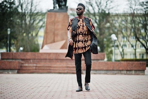 Homem africano elegante e bonito com roupa tradicional e boné preto em pé ao ar livre.