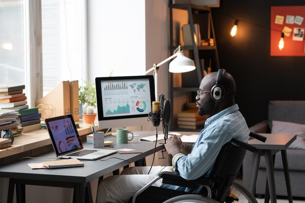 Homem africano em fones de ouvido sentado na cadeira de rodas na mesa em frente ao laptop com gráficos e falando ao microfone durante a conferência online