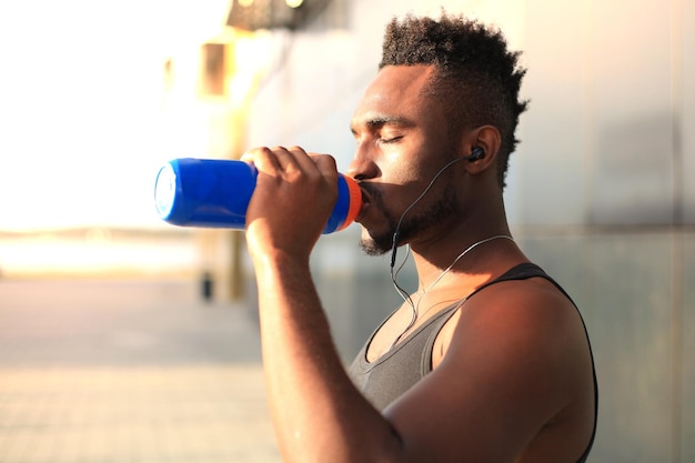 Homem africano em roupas esportivas, beber água enquanto está do lado de fora, ao pôr do sol ou nascer do sol. Corredor.
