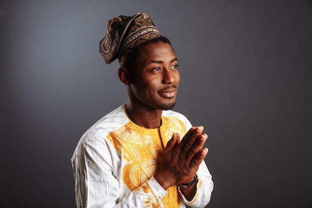 Homem africano em roupas nacionais e cocar tradicional Gele da Nigéria está rezando diante da parede cinza
