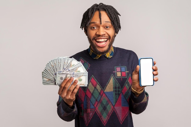 Homem africano extremamente feliz com dreadlocks satisfeito segurando telefone celular e fã de dólares nas mãos com sorriso dentuço, ganhando online. Tiro de estúdio interior isolado em fundo cinza