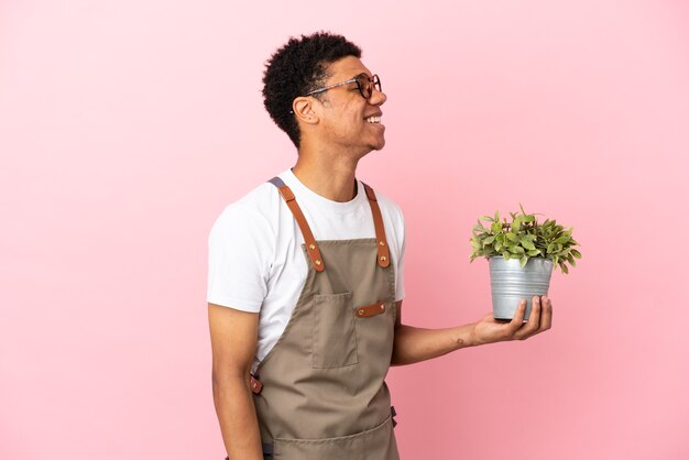 Homem africano jardineiro segurando uma planta isolada em um fundo rosa rindo em posição lateral