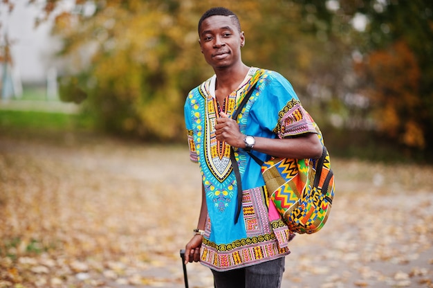 Homem Africano na camisa tradicional de África no outono park com mochila e mala. Viajante emigrante.