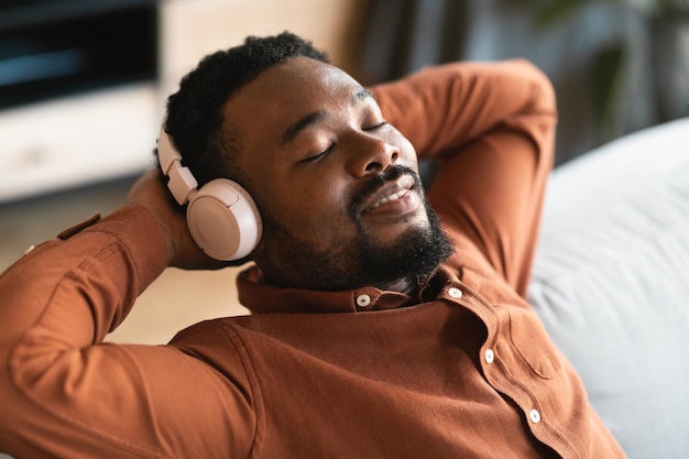 Homem africano ouvindo música usando fones de ouvido com os olhos fechados dentro de casa