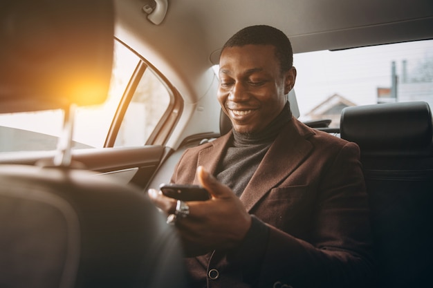 Foto homem africano que usa o smartphone ao sentar-se no assento traseiro no carro.