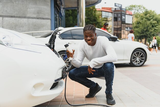 Homem africano segurando o cabo de carga disponível, em pé perto de um carro elétrico de luxo.