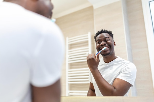 Homem africano sorridente com escova de dentes limpando os dentes e espelho no banheiro