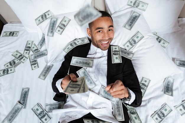 Foto homem africano sorridente de terno deitado na cama em um quarto de hotel com dinheiro caindo e olhando para a câmera. vista do topo