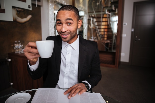 Homem africano sorridente de terno sentado com um diário e café no hotel