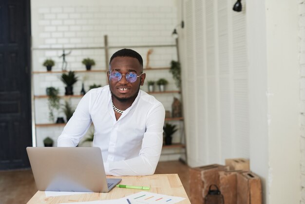 Homem africano sorridente, trabalhando em um escritório moderno, atrás de um laptop e olhando para a câmera