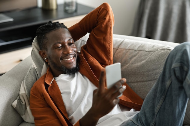 Homem afro-americano alegre assistindo vídeo no smartphone descansando e relaxando no sofá no interior da sala de estar