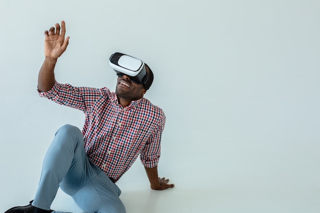 Homem afro-americano alegre e simpático testando óculos de realidade virtual enquanto está sentado no chão