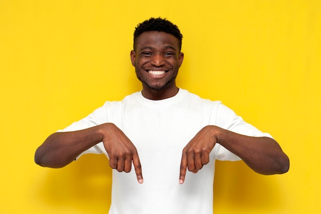 Homem afro-americano alegre em camisa branca mostra suas mãos para baixo em fundo isolado amarelo