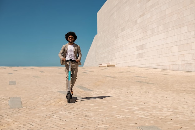 Homem afro-americano animado andando de scooter elétrica. Homem com cabelo encaracolado com óculos de sol, passar tempo ao ar livre. Tecnologia, conceito de atividade