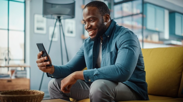 Homem afro-americano animado fazendo uma videochamada no smartphone enquanto está sentado em um sofá na sala de estar Homem feliz sorrindo em casa e falando com colegas e clientes pela Internet