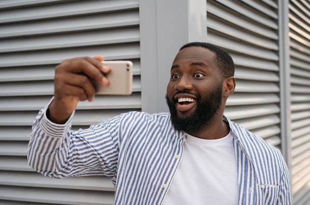 Homem afro-americano animado tomando selfie usando telefone móvel em pé na rua urbana. Blogueiro emocional influenciador filmando vídeo