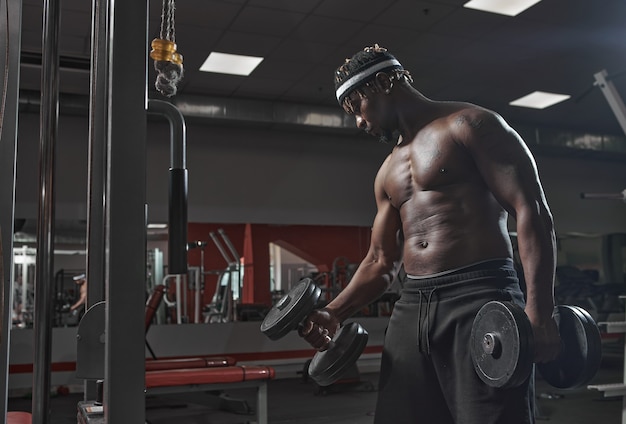 Homem afro-americano atlético se exercitando com halteres no treinamento de ginástica, perdendo peso e saudável