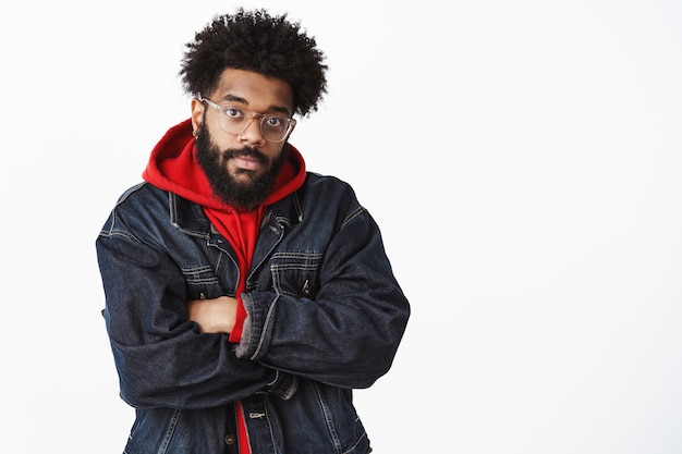 Homem afro-americano atraente e intenso com barba e penteado afro em jaqueta jeans sobre moletom com capuz e óculos cruzando os braços sobre o peito, sentindo frio e frio esperando por um amigo na rua