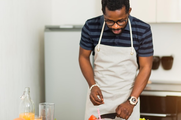 Homem afro-americano barbudo atraente com roupas casuais e avental, preparando o café da manhã com tomate, pepino, pimentão, berinjela, salada de folhas na cozinha. Comida saudável, conceito de comida caseira.