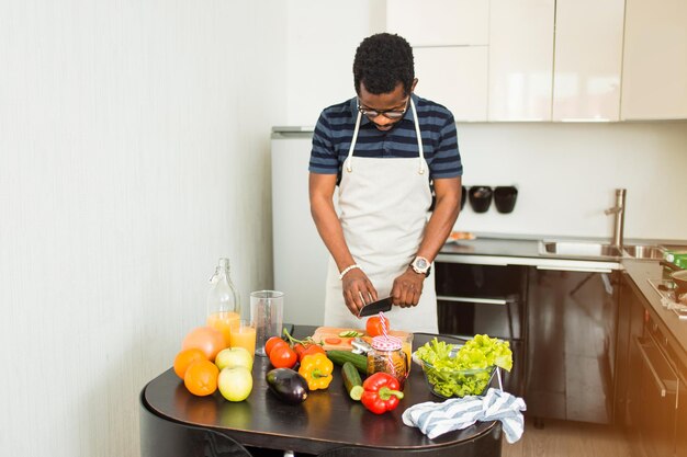 Homem afro-americano bonito cozinhar salada de vegetais, cortar tomates e pepinos na tábua, preparar o jantar em casa na cozinha. Comida saudável, conceito de cozinha.