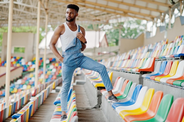 Homem afro-americano bonito de macacão jeans posado em cadeiras coloridas no estádio retrato de homem negro na moda